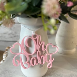 May include: A white ceramic vase with a pink "Oh Baby" sign hanging from it. The vase is filled with pink and white flowers.