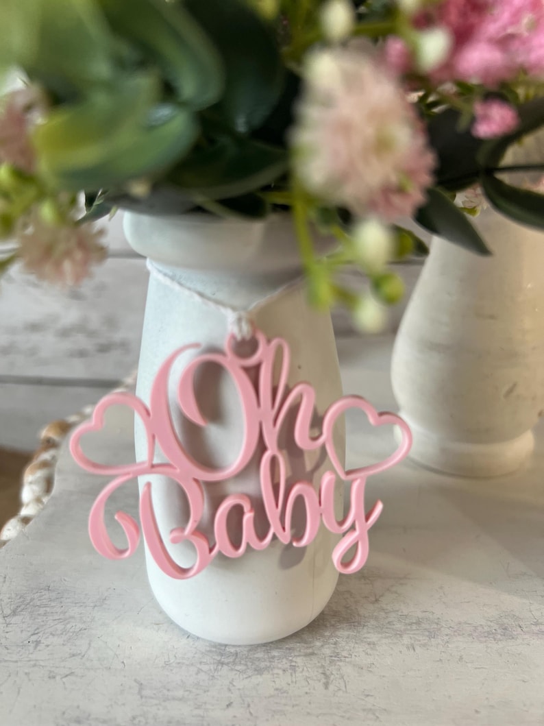 May include: A white ceramic vase with a pink "Oh Baby" sign hanging from it. The vase is filled with pink and white flowers.