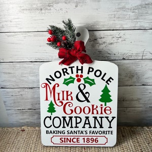 May include: White wooden sign with red and green Christmas decorations. The sign reads "North Pole Milk & Cookie Company Baking Santa's Favourite Since 1896".