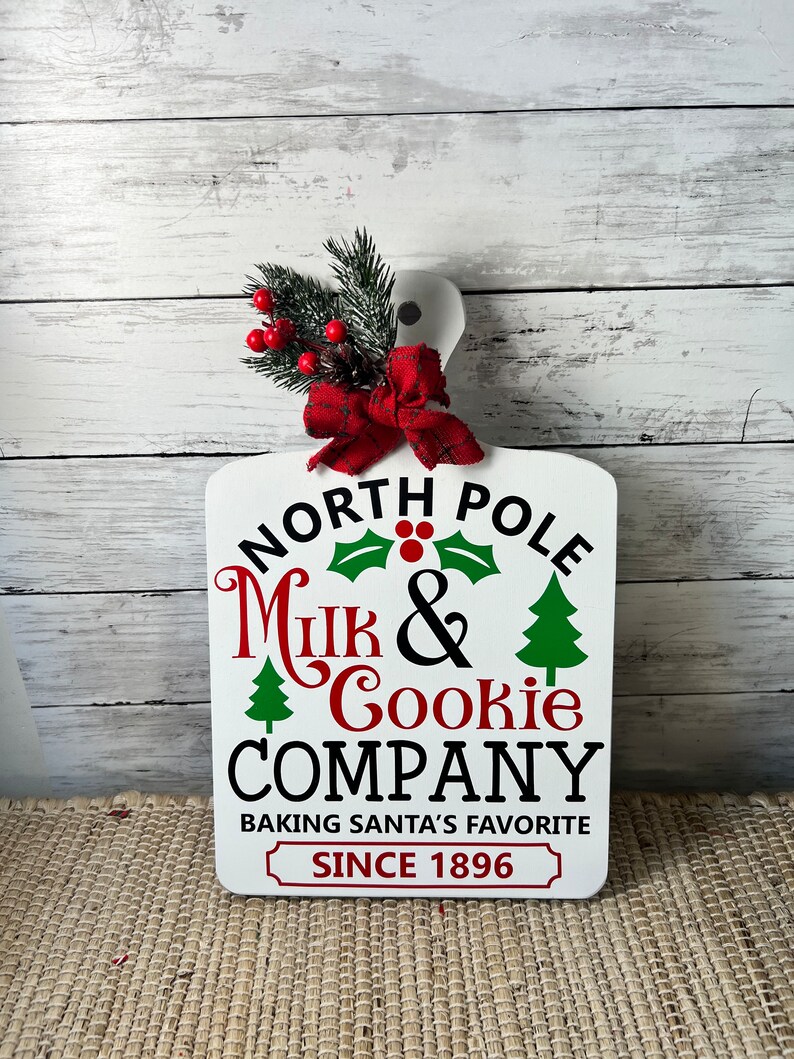 May include: White wooden sign with red and green Christmas decorations. The sign reads "North Pole Milk & Cookie Company Baking Santa's Favourite Since 1896".