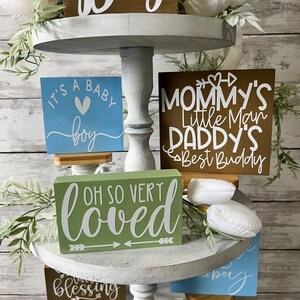 K&ouml;nnte beinhalten: Ein dreistufiges Tablett, dekoriert mit Holzschildchen mit den Aufschriften "It's a Baby Boy", "Oh So Very Loved", "Mommy's Little Man Daddy's Best Buddy" und "Blessing". Das Tablett ist au&szlig;erdem mit wei&szlig;en Blumen und Gr&uuml;npflanzen verziert.
