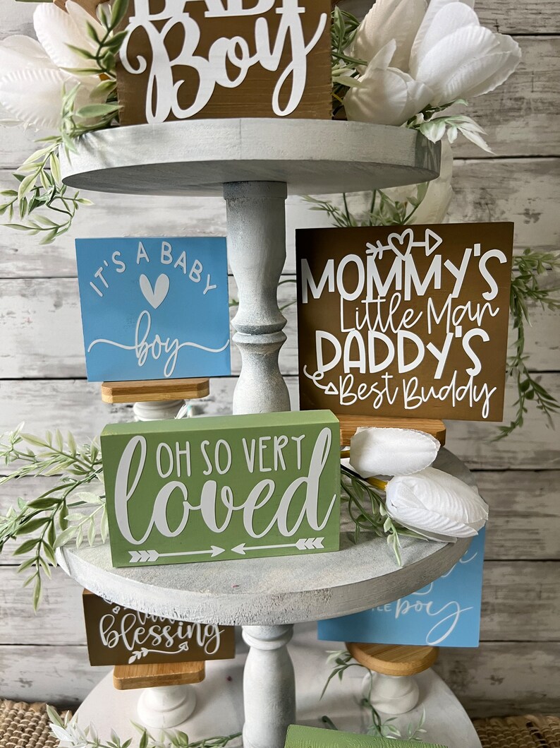 K&ouml;nnte beinhalten: Ein dreistufiges Tablett, dekoriert mit Holzschildchen mit den Aufschriften "It's a Baby Boy", "Oh So Very Loved", "Mommy's Little Man Daddy's Best Buddy" und "Blessing". Das Tablett ist au&szlig;erdem mit wei&szlig;en Blumen und Gr&uuml;npflanzen verziert.