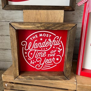May include: A wooden framed sign with a red background and white text that reads "The Most Wonderful Time of the Year".