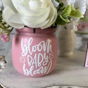 May include: A pink ceramic vase with a white floral design and the text "bloom baby bloom" in white lettering. The vase is filled with white and pink flowers.