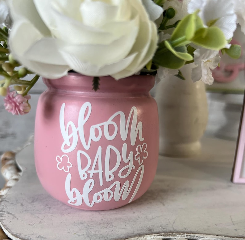 May include: A pink ceramic vase with a white floral design and the text "bloom baby bloom" in white lettering. The vase is filled with white and pink flowers.