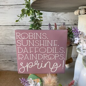 Pu&ograve; includere: Un cartello di legno rosa con lettere bianche che recita "ROBINS. SUNSHINE. DAFFODILS. RAINDROPS. spring!" Il cartello &egrave; appoggiato su un vassoio a pi&ugrave; livelli con verde e fiori.