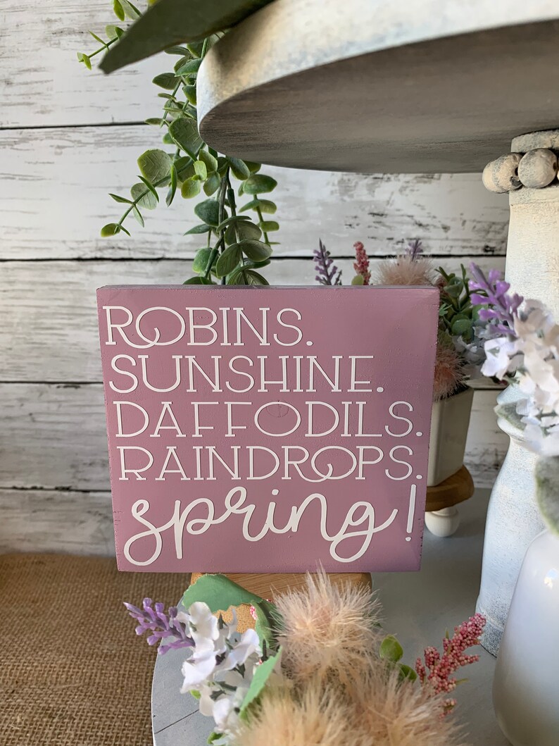 Pu&ograve; includere: Un cartello di legno rosa con lettere bianche che recita "ROBINS. SUNSHINE. DAFFODILS. RAINDROPS. spring!" Il cartello &egrave; appoggiato su un vassoio a pi&ugrave; livelli con verde e fiori.