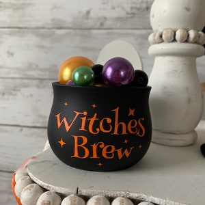 May include: Black cauldron with the text "Witches Brew" in orange lettering, filled with colorful beads, on a white shelf with a white beaded base.