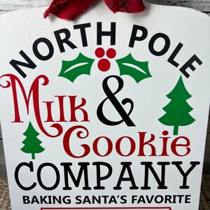 May include: White wooden sign with black text that reads "North Pole Milk & Cookie Company Baking Santa's Favourite Since 1896". The sign has a red bow at the top and green and red Christmas decorations.
