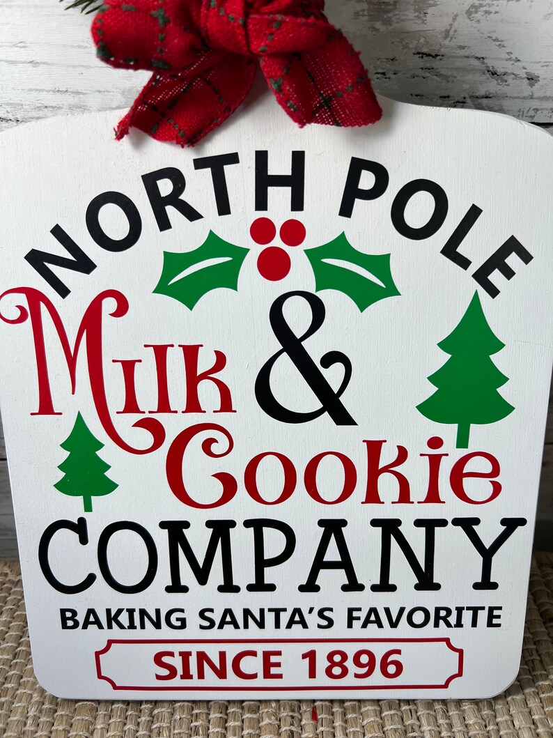 May include: White wooden sign with black text that reads "North Pole Milk & Cookie Company Baking Santa's Favourite Since 1896". The sign has a red bow at the top and green and red Christmas decorations.