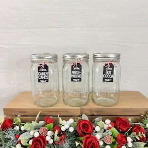 May include: Three clear glass jars with silver lids and red and white twine tied around the lids. Each jar has a black tag with white text. The tags read "Candy Canes", "Marshmallows", and "Hot Cocoa". The jars are sitting on a wooden surface with a red and white garland in front of them.