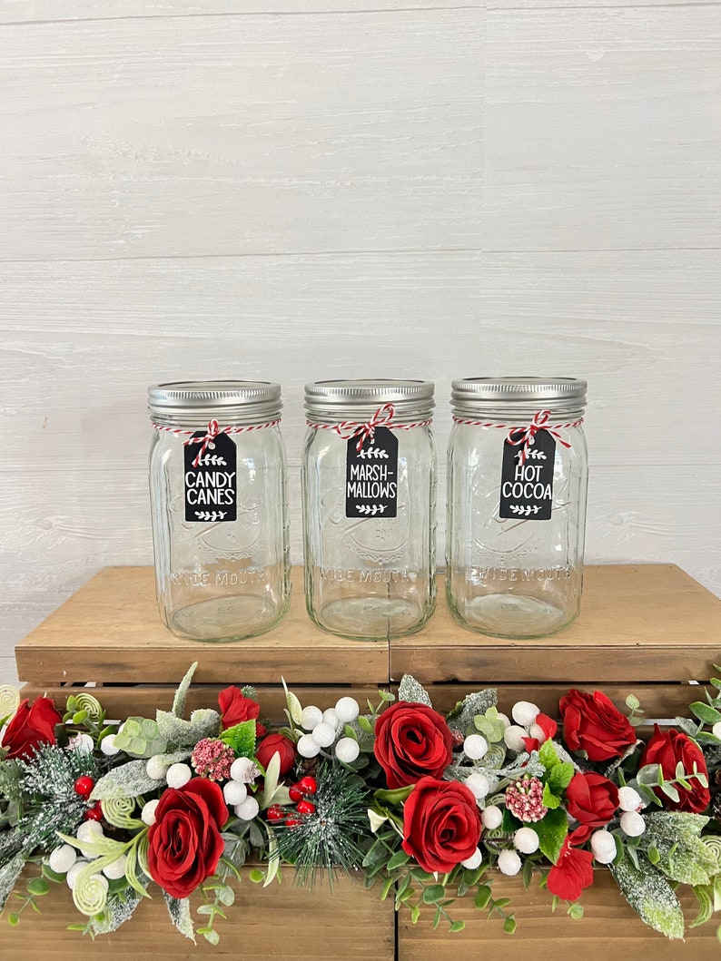 May include: Three clear glass jars with silver lids and red and white twine tied around the lids. Each jar has a black tag with white text. The tags read "Candy Canes", "Marshmallows", and "Hot Cocoa". The jars are sitting on a wooden surface with a red and white garland in front of them.