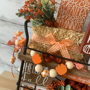May include: A tiered metal stand decorated for fall with a hay bale, orange pumpkins, fall-themed signs, and a garland of wooden beads and orange pumpkin charms. The signs read "Hello Fall" and "Bonfire, Pumpkin Spice, Sweaters, Smores, Flannels".
