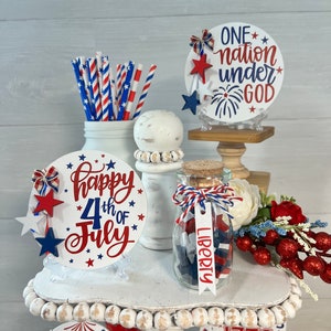 May include: A tiered tray decorated for the 4th of July with red, white, and blue decorations. The tray features a sign that says "Happy 4th of July", a jar filled with red, white, and blue stars, and a sign that says "One nation under God".