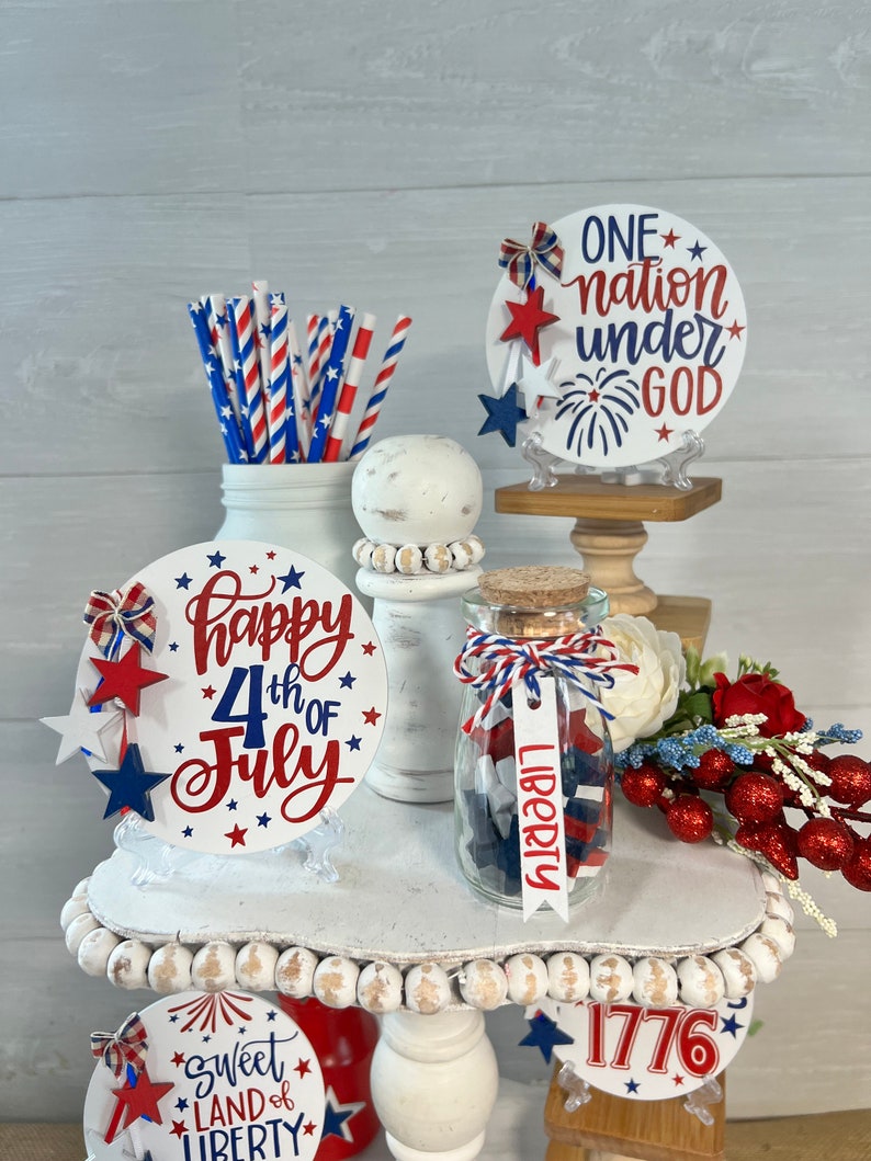 May include: A tiered tray decorated for the 4th of July with red, white, and blue decorations. The tray features a sign that says "Happy 4th of July", a jar filled with red, white, and blue stars, and a sign that says "One nation under God".