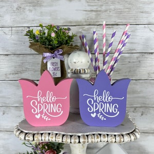 K&ouml;nnte beinhalten: Zwei h&ouml;lzerne tulpenf&ouml;rmige Schilder mit dem Text "hello spring" in wei&szlig;en Buchstaben. Die Schilder sind in rosa und lila lackiert und stehen auf einem wei&szlig;en, mehrstufigen St&auml;nder. Auf der obersten Stufe des St&auml;nders befindet sich ein kleiner Blumenstrau&szlig; in einer mit Sackleinen umwickelten Vase mit einem Schild, auf dem "fresh flowers 25c" steht.