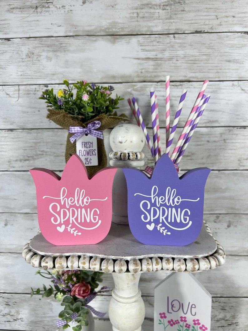 K&ouml;nnte beinhalten: Zwei h&ouml;lzerne tulpenf&ouml;rmige Schilder mit dem Text "hello spring" in wei&szlig;en Buchstaben. Die Schilder sind in rosa und lila lackiert und stehen auf einem wei&szlig;en, mehrstufigen St&auml;nder. Auf der obersten Stufe des St&auml;nders befindet sich ein kleiner Blumenstrau&szlig; in einer mit Sackleinen umwickelten Vase mit einem Schild, auf dem "fresh flowers 25c" steht.