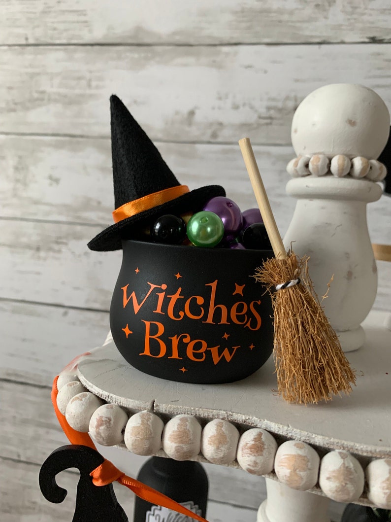 May include: Black cauldron with orange text that reads "Witches Brew" and a black witch hat with an orange ribbon. The cauldron is filled with colorful beads and a small brown broom is next to it.