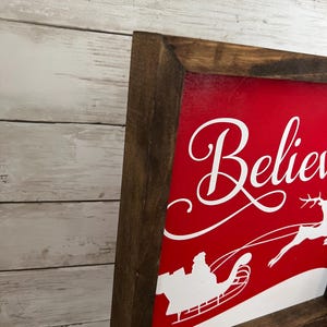 May include: A red wooden sign with a white silhouette of Santa Claus in his sleigh flying through the air. The sign says "Believe" in white cursive lettering.