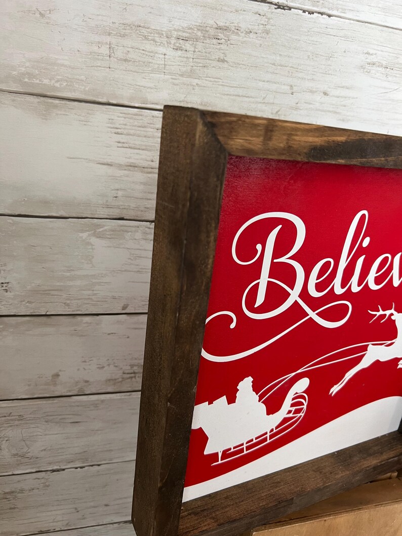 May include: A red wooden sign with a white silhouette of Santa Claus in his sleigh flying through the air. The sign says "Believe" in white cursive lettering.