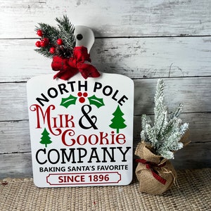 May include: White wooden chopping board with the text "North Pole Milk & Cookie Company Baking Santa's Favourite Since 1896". The board is decorated with a red bow and a sprig of greenery with red berries.