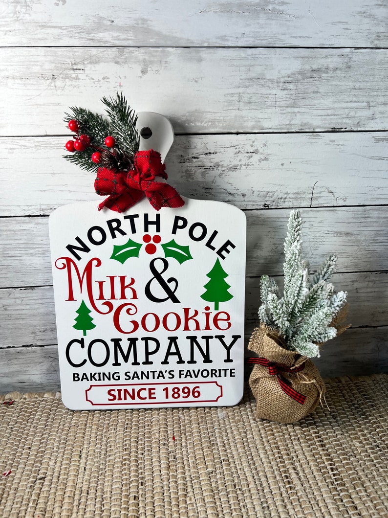 May include: White wooden chopping board with the text "North Pole Milk & Cookie Company Baking Santa's Favourite Since 1896". The board is decorated with a red bow and a sprig of greenery with red berries.