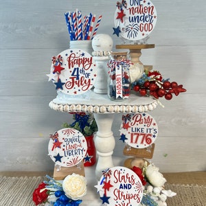 May include: A tiered white cake stand decorated with red, white, and blue patriotic themed round signs. The signs feature phrases like "Happy 4th of July", "Sweet Land of Liberty", "Stars and Stripes", "Party Like It's 1776", and "One Nation Under God".