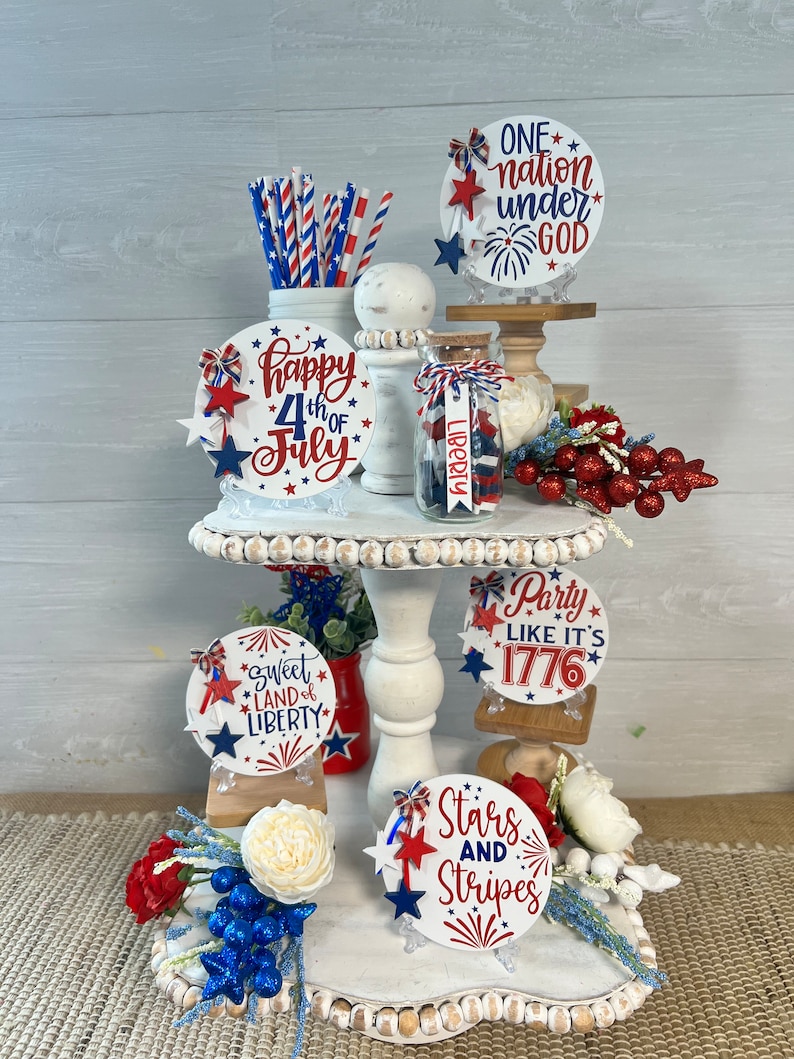 May include: A tiered white cake stand decorated with red, white, and blue patriotic themed round signs. The signs feature phrases like "Happy 4th of July", "Sweet Land of Liberty", "Stars and Stripes", "Party Like It's 1776", and "One Nation Under God".