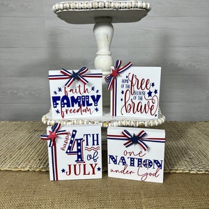 May include: Four wooden blocks with red, white, and blue patriotic decorations and text. The blocks read "faith family freedom", "home of the free because of the brave", "happy 4th of july", and "one nation under god".