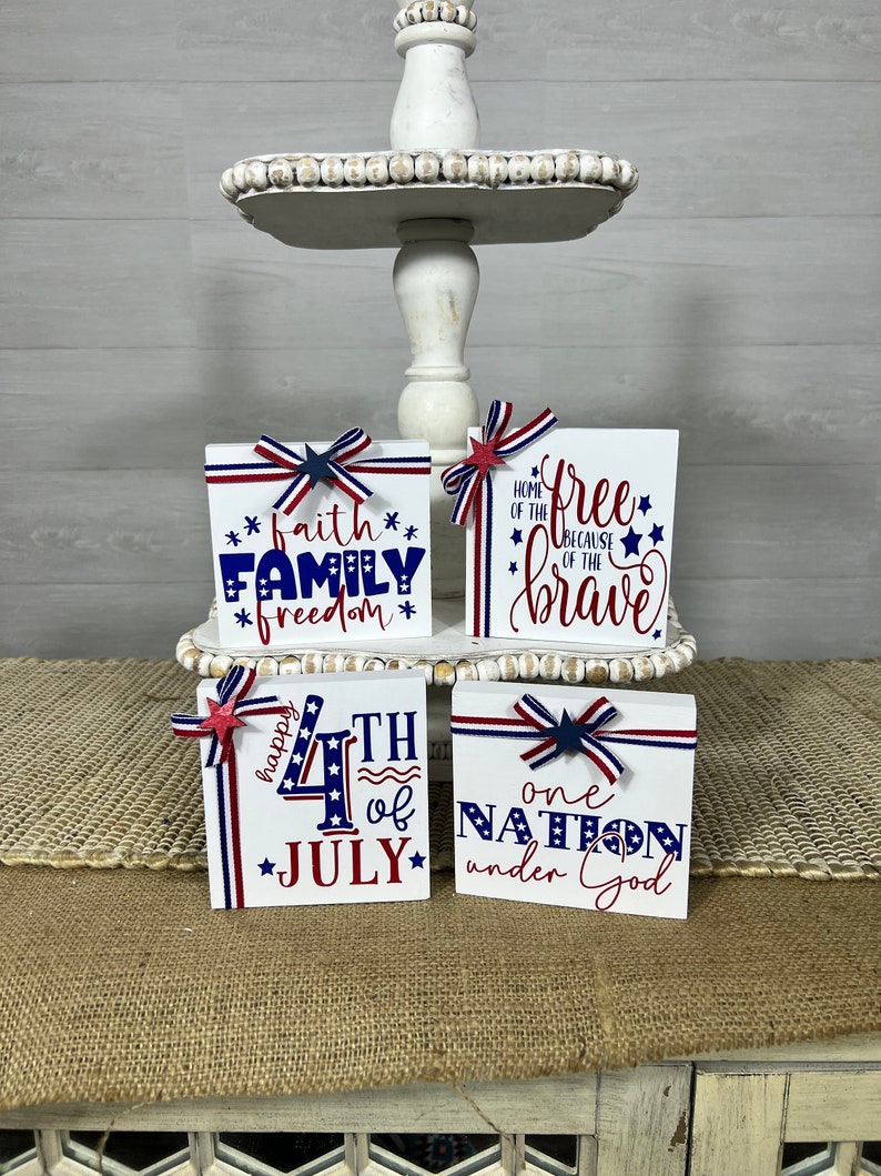 May include: Four wooden blocks with red, white, and blue patriotic decorations and text. The blocks read "faith family freedom", "home of the free because of the brave", "happy 4th of july", and "one nation under god".