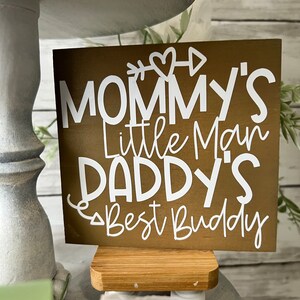 K&ouml;nnte beinhalten: Ein braunes Holzschild mit wei&szlig;em Text, der "Mommy's Little Man Daddy's Best Buddy" lautet, mit einem Herz- und Pfeil-Motiv. Das Schild steht auf einem kleinen Holzst&auml;nder.