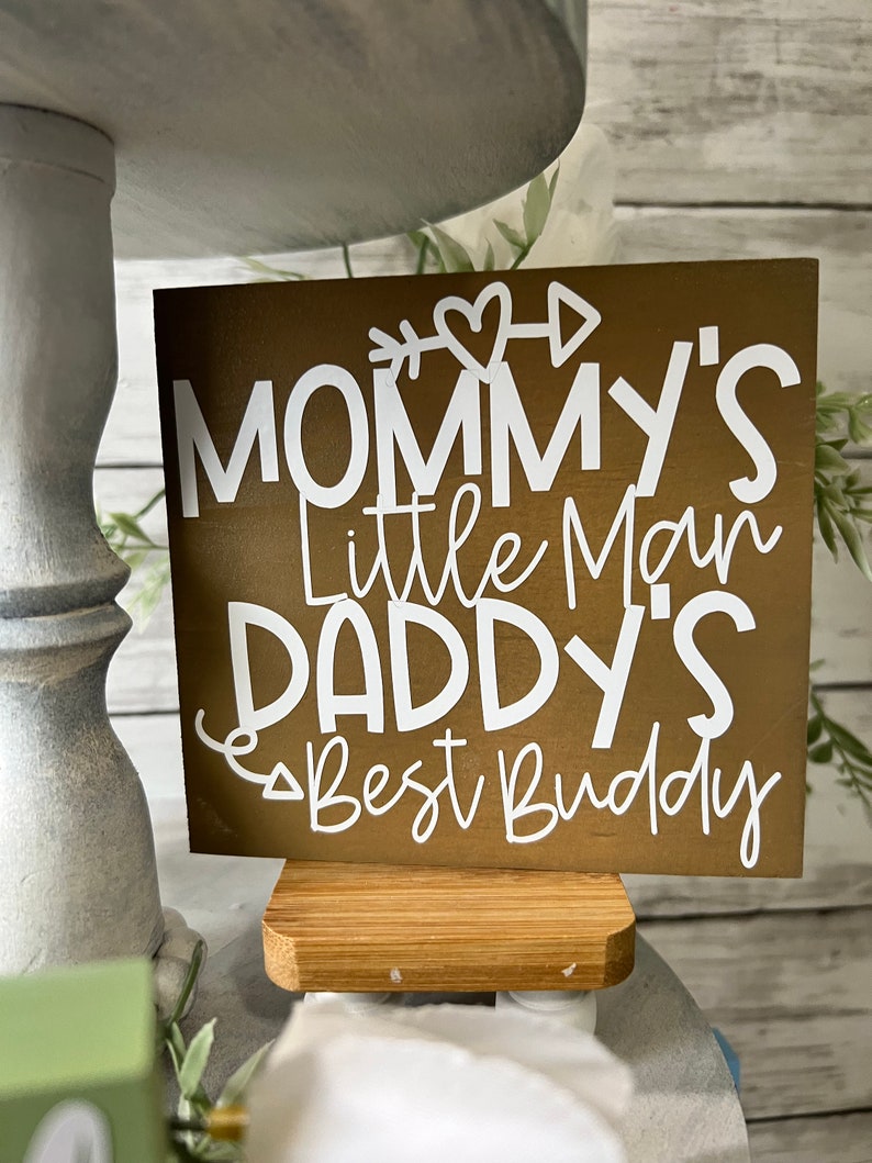 K&ouml;nnte beinhalten: Ein braunes Holzschild mit wei&szlig;em Text, der "Mommy's Little Man Daddy's Best Buddy" lautet, mit einem Herz- und Pfeil-Motiv. Das Schild steht auf einem kleinen Holzst&auml;nder.