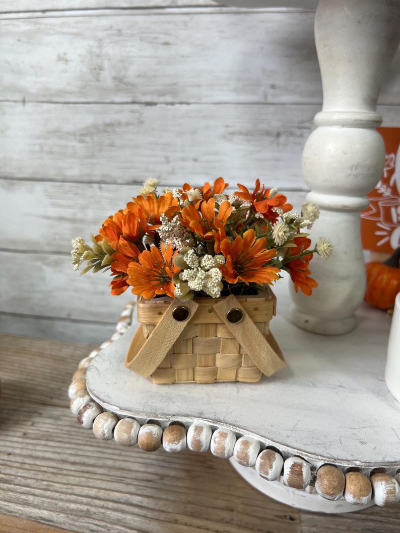 May include: A small woven basket with orange and white flowers. The basket has two leather handles and is sitting on a white tiered cake stand with a beaded edge.
