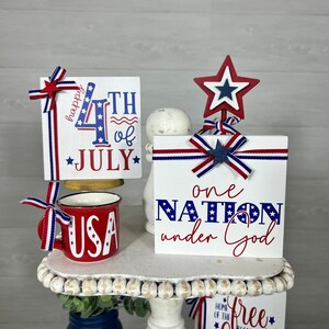 May include: A tiered white stand decorated with red, white, and blue patriotic themed wooden signs. The signs read "Happy 4th of July", "One Nation Under God", and "Free Because of the Brave".