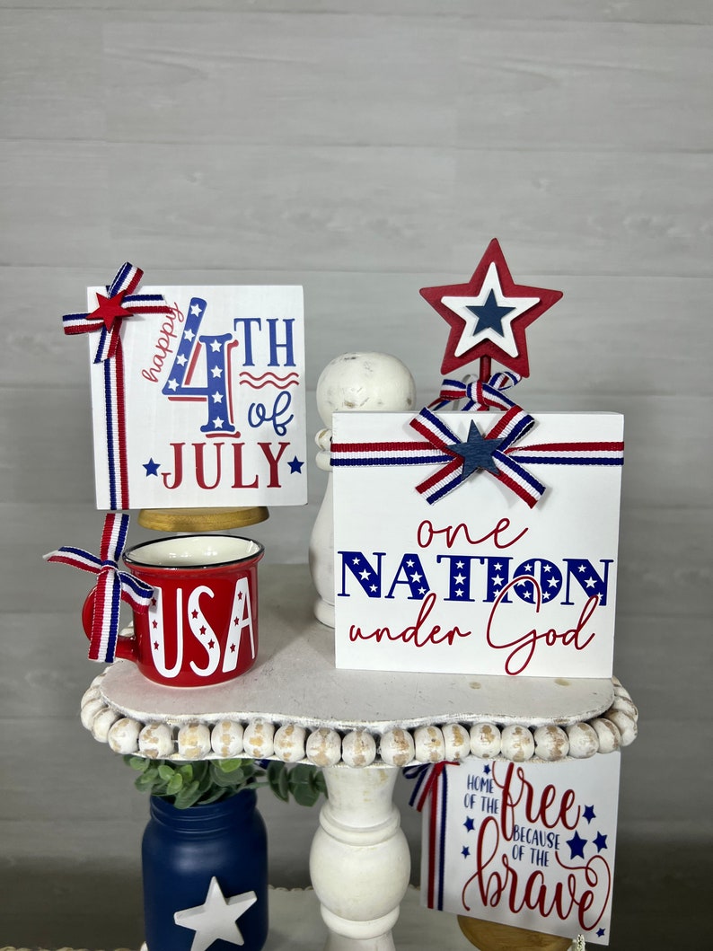 May include: A tiered white stand decorated with red, white, and blue patriotic themed wooden signs. The signs read "Happy 4th of July", "One Nation Under God", and "Free Because of the Brave".