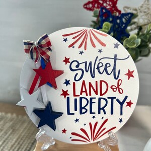 May include: White round wooden sign with the text "Sweet Land of Liberty" in blue and red lettering. The sign is decorated with red, white, and blue stars and fireworks. The sign is on a small wooden stand.