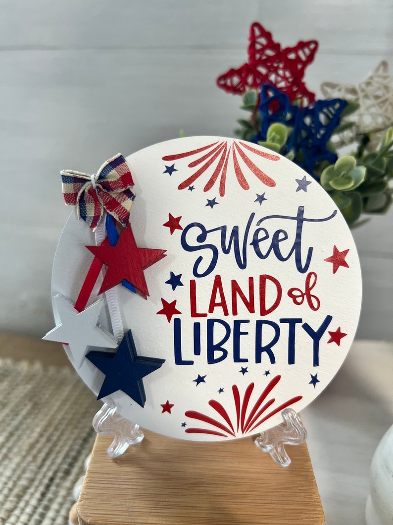 May include: White round wooden sign with the text "Sweet Land of Liberty" in blue and red lettering. The sign is decorated with red, white, and blue stars and fireworks. The sign is on a small wooden stand.