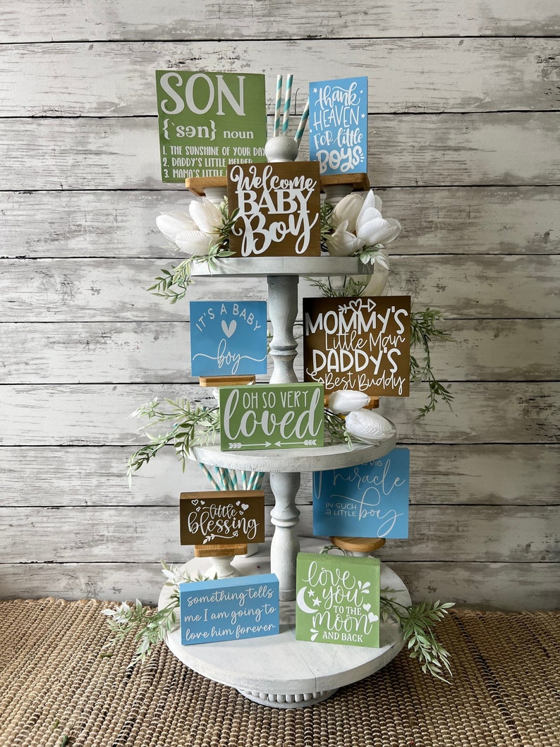 K&ouml;nnte beinhalten: Ein dreistufiges Tablett, das mit verschiedenen Holzschildern f&uuml;r eine Babyparty dekoriert ist. Die Schilder zeigen S&auml;tze wie "Willkommen Baby Boy", "Es ist ein Baby Boy", "Mamas kleiner Mann", "Papas bester Freund", "So sehr geliebt", "Kleiner Segen", "Wunderkind", "Ich liebe dich bis zum Mond und zur&uuml;ck" und "Etwas sagt mir, dass ich ihn f&uuml;r immer lieben werde".
