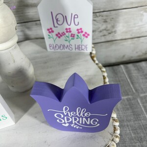 K&ouml;nnte beinhalten: Lilafarbener, tulpenf&ouml;rmiger Holzschild mit dem Text "hello spring" in wei&szlig;er Farbe. Das Schild steht auf einem wei&szlig;en Regal mit einem wei&szlig;en, hausf&ouml;rmigen Holzschild, auf dem "love blooms here" in lila Farbe steht.