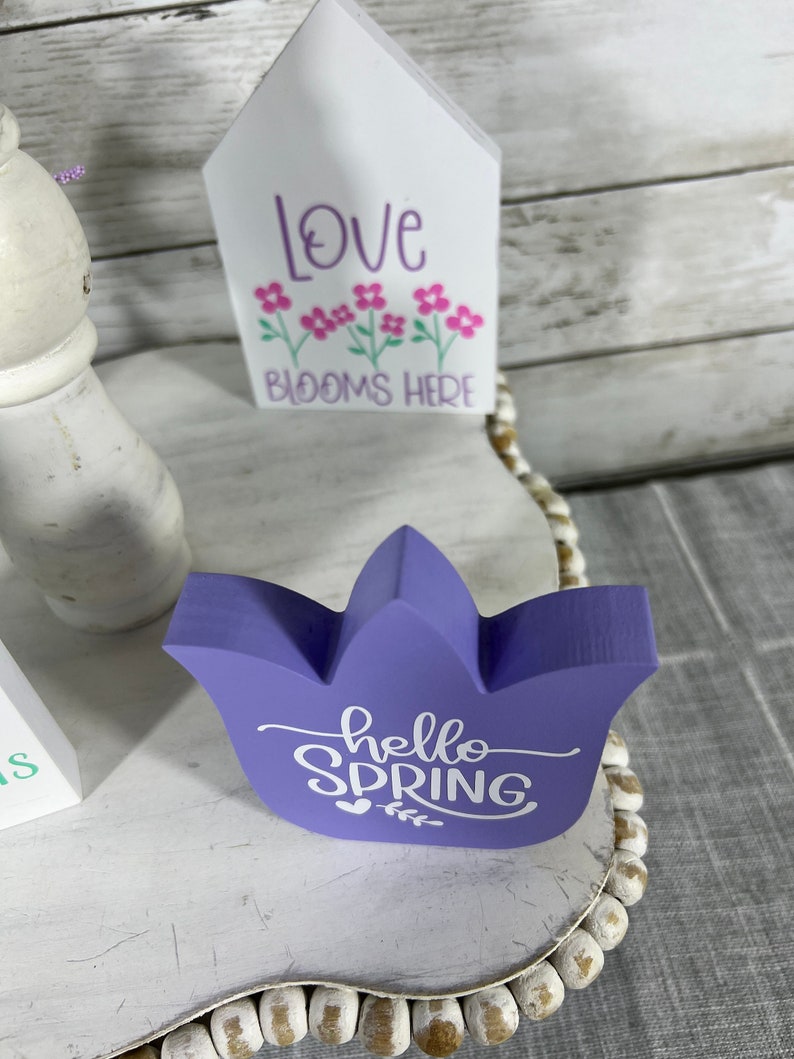K&ouml;nnte beinhalten: Lilafarbener, tulpenf&ouml;rmiger Holzschild mit dem Text "hello spring" in wei&szlig;er Farbe. Das Schild steht auf einem wei&szlig;en Regal mit einem wei&szlig;en, hausf&ouml;rmigen Holzschild, auf dem "love blooms here" in lila Farbe steht.