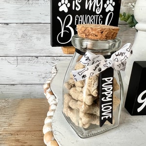 May include: A glass jar with a cork lid filled with dog treats. The jar is tied with a white ribbon with the word "Woof!" printed on it. A black tag with the text "Puppy Love" is attached to the ribbon. The jar is sitting on a white tiered tray with brown beads around the edge. A black sign with white text reads "RESCUED is my FAVORITE" with paw prints.