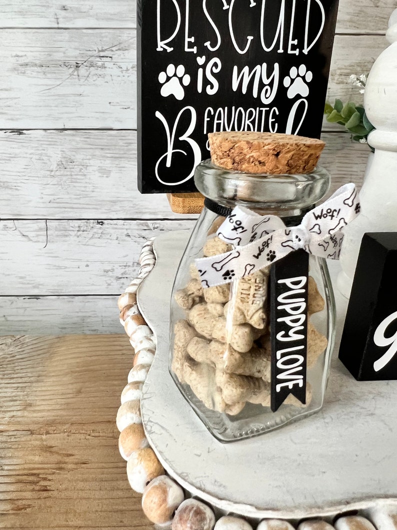 May include: A glass jar with a cork lid filled with dog treats. The jar is tied with a white ribbon with the word "Woof!" printed on it. A black tag with the text "Puppy Love" is attached to the ribbon. The jar is sitting on a white tiered tray with brown beads around the edge. A black sign with white text reads "RESCUED is my FAVORITE" with paw prints.