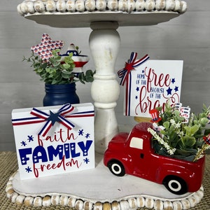 May include: A tiered tray decorated with red, white, and blue patriotic themed decor. The top tier features a red mug with the word "USA" on it, a wooden sign that says "nation under God", and white beads. The middle tier features a blue mason jar with greenery, a red, white, and blue star, and a wooden sign that says "home of the free because of the brave". The bottom tier features a red truck with a white bed, a wooden sign that says "faith family freedom", and white beads.