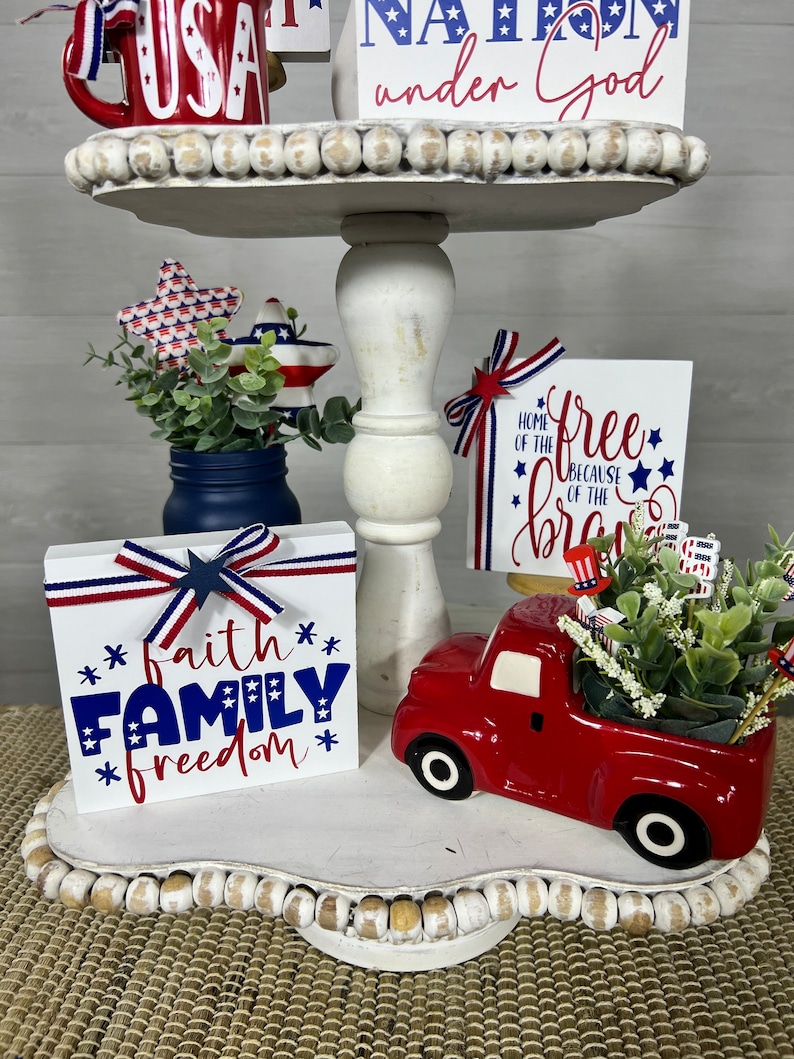 May include: A tiered tray decorated with red, white, and blue patriotic themed decor. The top tier features a red mug with the word "USA" on it, a wooden sign that says "nation under God", and white beads. The middle tier features a blue mason jar with greenery, a red, white, and blue star, and a wooden sign that says "home of the free because of the brave". The bottom tier features a red truck with a white bed, a wooden sign that says "faith family freedom", and white beads.