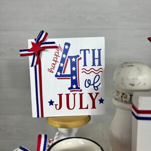 May include: A white card with red, white, and blue stars and stripes design. The card says "happy 4th of JULY" with a red star on each side of the word "JULY". The card is on a small wooden stand.