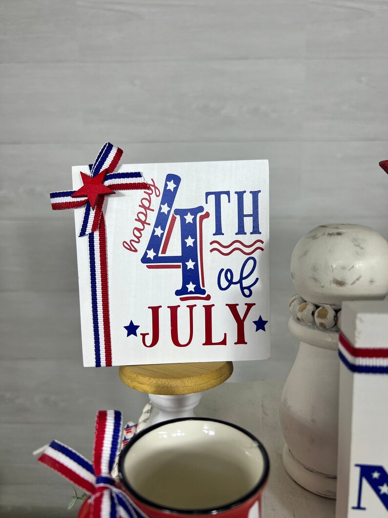 May include: A white card with red, white, and blue stars and stripes design. The card says "happy 4th of JULY" with a red star on each side of the word "JULY". The card is on a small wooden stand.