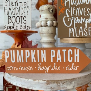 May include: A set of four wooden signs with fall-themed text. The signs are painted in various colors, including brown, orange, and white. The text on the signs reads: "pumpkins bonfires flannel hayrides boots apple cider", "Autumn leaves pumpkin please", "PUMPKIN PATCH corn maze hayrides cider", and "FALL".