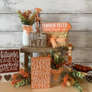 May include: A fall-themed tiered tray display with various wooden signs featuring autumn-inspired phrases like "Pumpkin Patch", "Hello Fall", "Autumn Leaves & Pumpkins Please", and "Bonfires Pumpkin Spice Sweaters Smores & Flannels". The tray is decorated with faux greenery, a small wagon, and a hay bale.