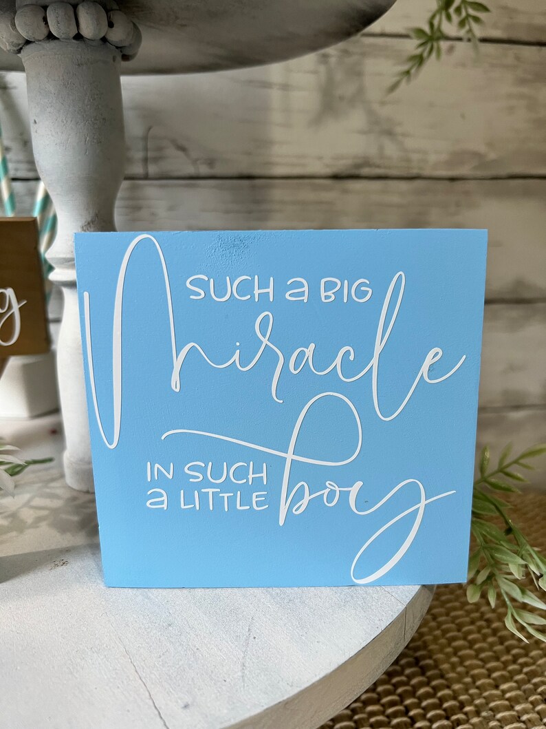 K&ouml;nnte beinhalten: Ein hellblaues Holzschild mit wei&szlig;er Aufschrift, die lautet "Such a big miracle in such a little boy".