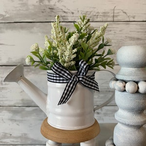 May include: A white metal watering can filled with white and green faux flowers, accented with a black and white checkered bow. The watering can sits on a small wooden stand with white legs. Decorative home decor.
